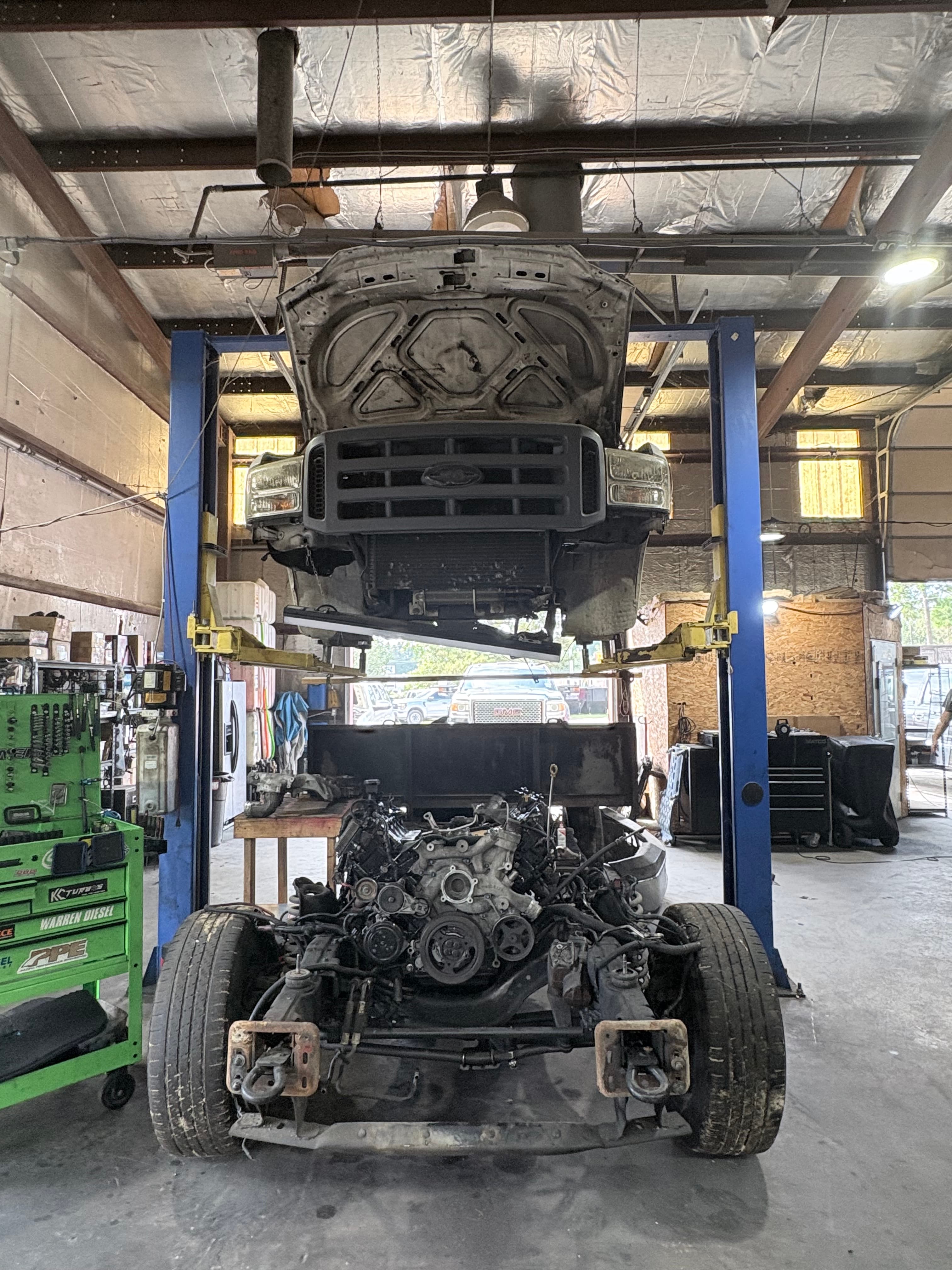 Ford truck body lifted on a blue hoist above its exposed engine and chassis.