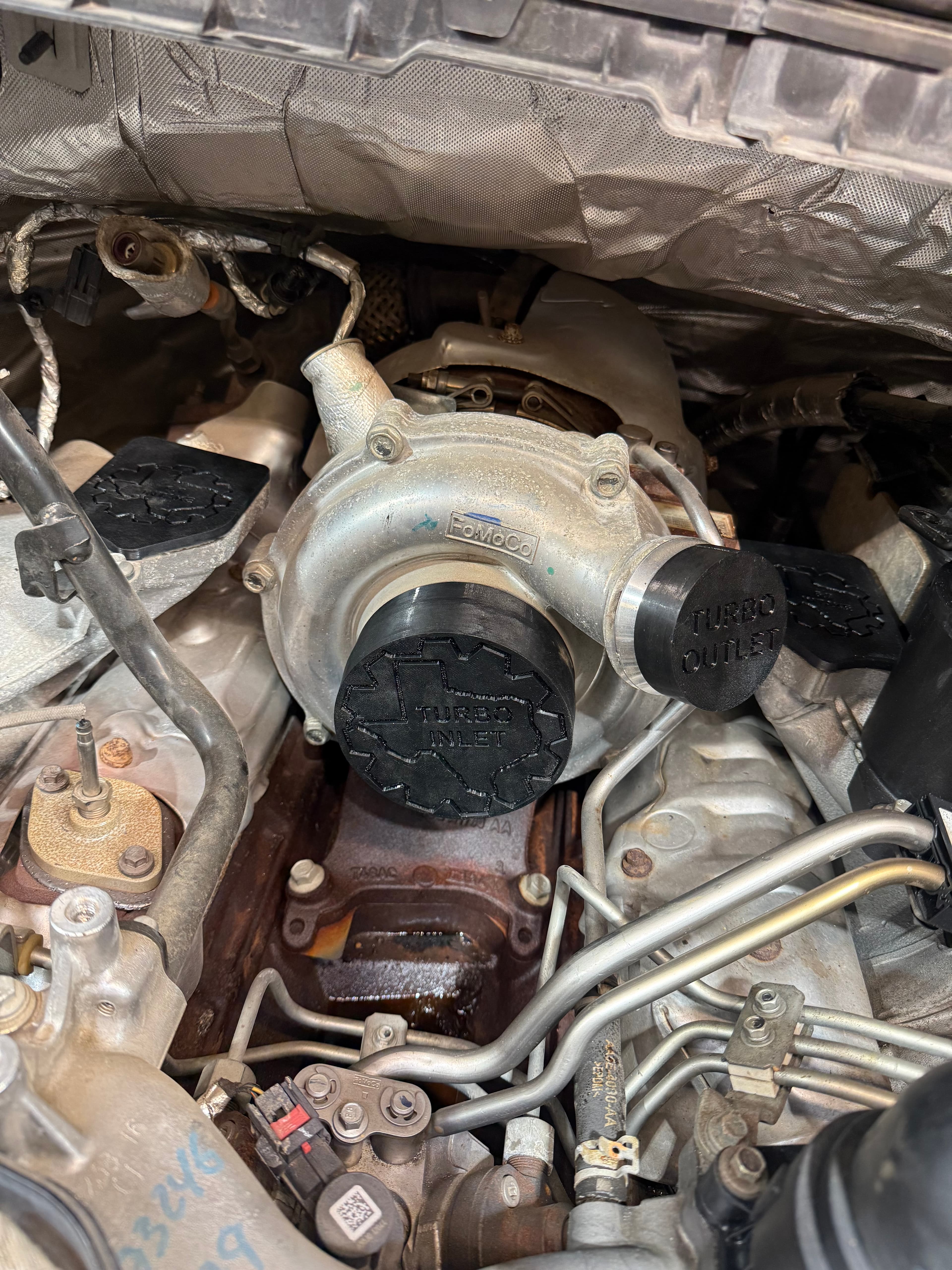 FoMoCo turbocharger with black inlet and outlet caps installed in a vehicle's engine bay.