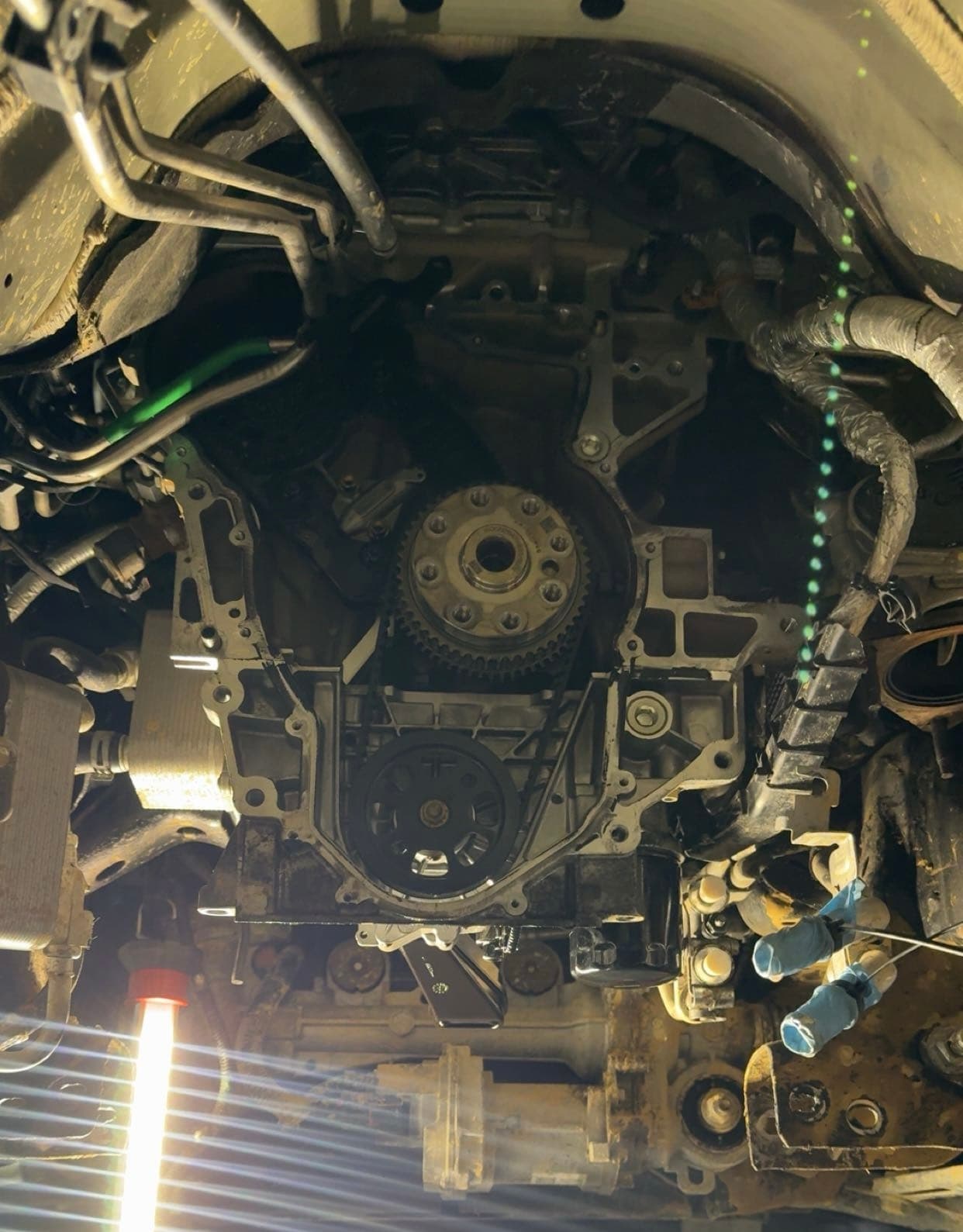 Exposed car engine showing timing gears and belt, illuminated by a bright work light from below.